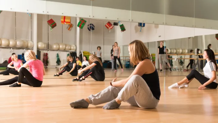 Kobiety uczestniczące w zajęciach tańca intuicyjnegZdjęcie przedstawia grupę kobiet uczestniczących w warsztatach tańca intuicyjnego. Panie siedzą na drewnianej podłodze w przestronnej sali z lustrami, wykonując ćwiczenia rozciągające i oswajające z ciałem. Atmosfera jest spokojna, wspierająca i skupiona na doświadczaniu siebie poprzez rucho w jasnej sali z lustrami