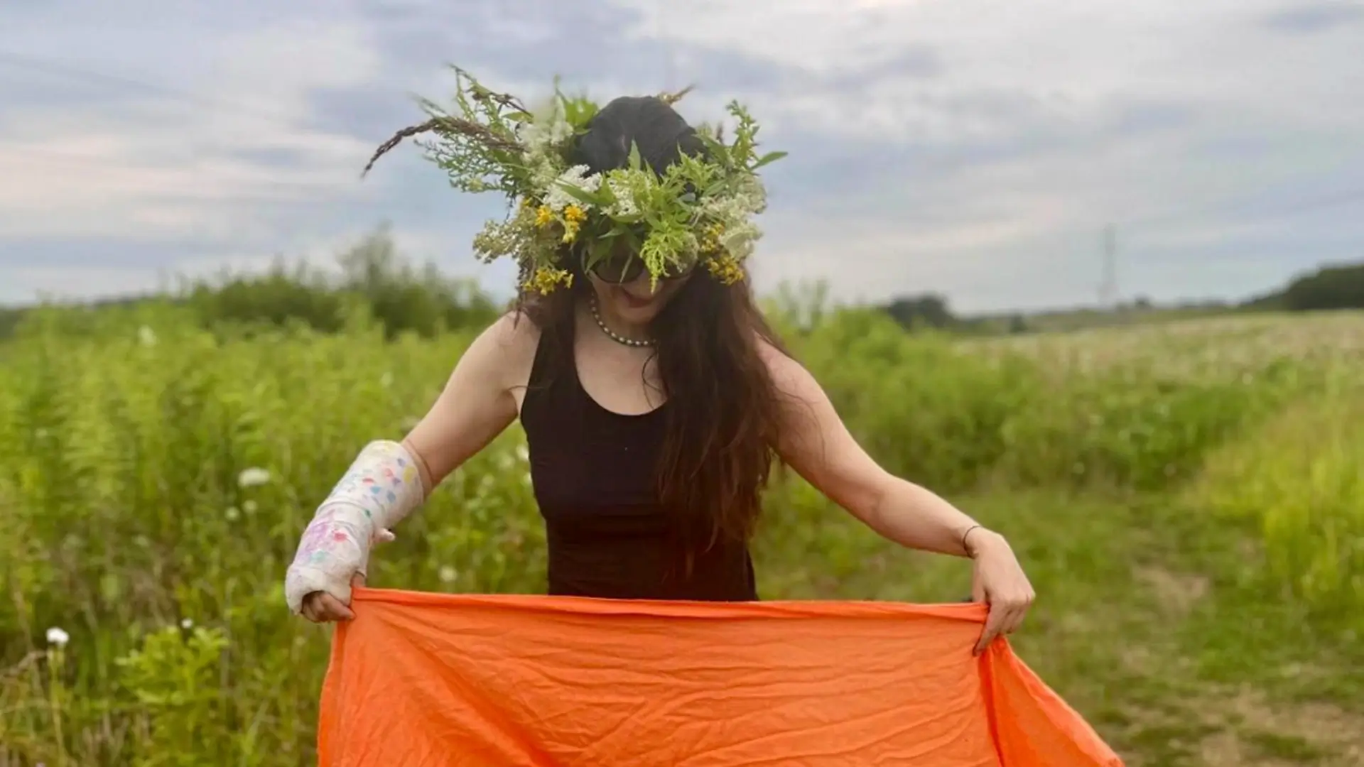 Kobieta w czarnej sukience i wianku z polnych kwiatów, z założonym gipsem na lewej ręce, stoi na łące i trzyma pomarańczową chustę, spoglądając w dół.